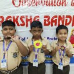 Three schoolboys in beige uniforms proudly display their medals and ribbons in front of a raksha bandhan event banner with festive colors